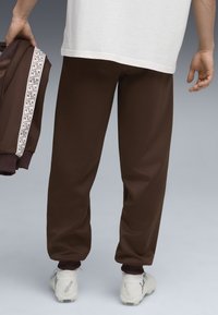 Brune sweatpants med ribbede manchetter, lavet af et glat materiale, parret med hvide sneakers. En matchende jakke draperet over armen har et mønstret stribe.