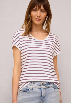 Donna che indossa una camicia bianca a maniche corte con strisce orizzontali viola e jeans di denim azzurro chiaro su uno sfondo semplice.