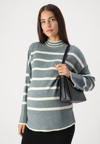 Hellblau gerippter Pullover mit elfenbeinfarbenen Streifen und hohem Stehkragen. Das Model hält eine schwarze, strukturiertes Handtasche mit einem kurzen Trageriemen.