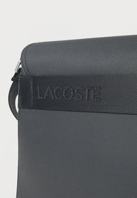 Κοντινό πλάνο μαύρης υφασμάτινης τσάντας με ανάγλυφο λογότυπο Lacoste σε οριζόντια λωρίδα κοντά στην πάνω άκρη.