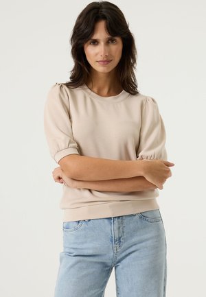 Sweatshirt beige à manches bouffantes et poignets côtelés, doté d'un col rond et d'un tissu texturé, associé à un jean bleu clair.