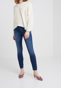 Bílý pletený svetr s výstřihem do V a otevřeným vzorem, spárovaný s tmavě modrými úzkými džíny a vzorovanými, nízkými podpatky.