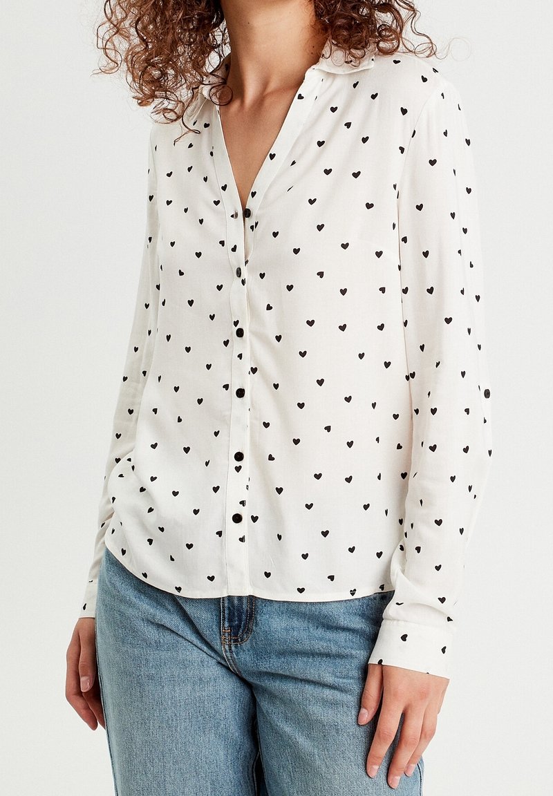 Chemise blanche à boutons avec petits motifs de cœurs noirs, portée avec un jean bleu clair, montrée du cou aux hanches.