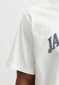 Flødefarvet bomuld T-shirt med løs pasform, der har et grafisk tryk af ordet "JA" i mørkeblå på brystet.