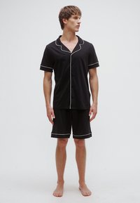 Σετ πυτζάμας με μαύρο κοντομάνικο, κατασκευασμένο από απαλό ύφασμα, με λευκή τρέσα κατά μήκος του γιακά, των μανσετών και των σορτς. Απλός σχεδιασμός με κουμπιά μπροστά.