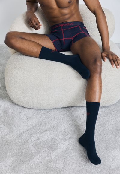 Marineblauwe boxershort met een rood ruitpatroon en het logo van Emporio Armani, gecombineerd met bijpassende marineblauwe crew sokken, model zittend op een luxe beige stoel.