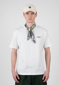 Jeune homme portant un T-shirt blanc à fines rayures, un pantalon sombre, une casquette beige et un foulard à motifs autour du cou, sur un fond uni.