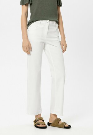 Frau trägt gerade geschnittene weiße Hose, olivgrünes kurzärmeliges Oberteil und olivgrüne Sandalen mit Metallnieten und Schnallen, steht vor weißem Hintergrund.