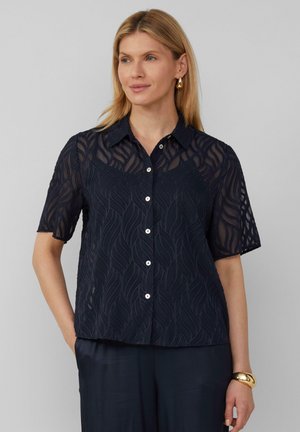 Chemise courte à manches courtes de couleur marine avec un motif en vagues texturé, boutonnée à l'avant et dotée d'un col classique. Le tissu semble léger et semi-transparent.