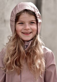 Otrok, ki nosi svetlo roza nepremočljivo jakno z oprijetim kapuco. Lasje so dolgi in valoviti, nasmeh pa razkriva zobe.