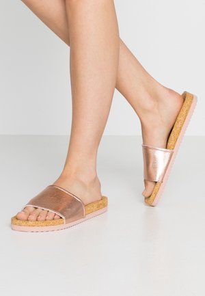 Nagie nogi w różowo-złotych klapkach z korkową wkładką i różowymi podeszwami na białym tle.