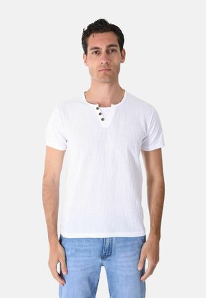 Uomo in piedi di fronte, indossa una maglietta henley bianca a maniche corte con tre bottoni e jeans azzurri chiari.