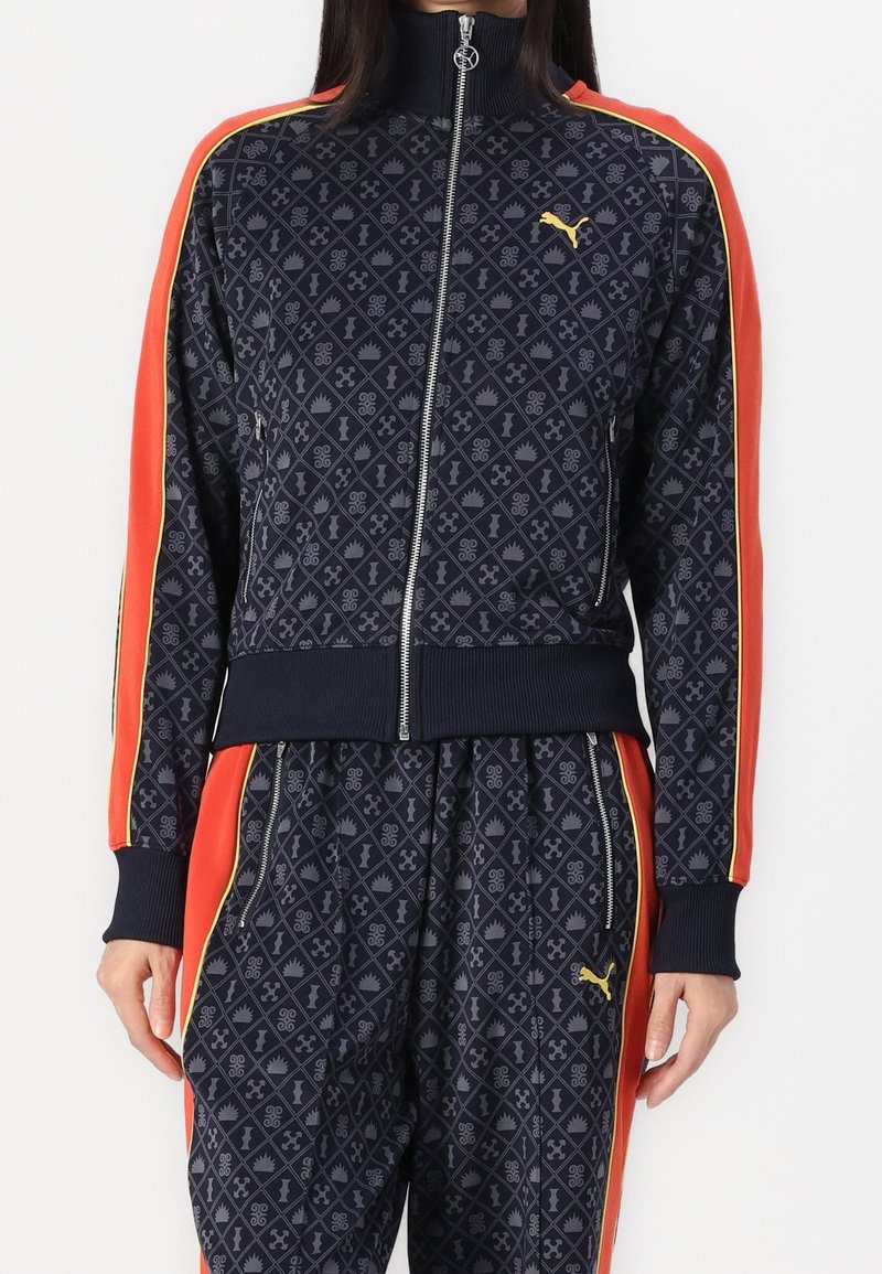 Femme portant un survêtement Puma bleu marine à motifs avec des rayures orange et jaunes sur les manches et le pantalon, veste à fermeture éclair frontale et poches zippées.