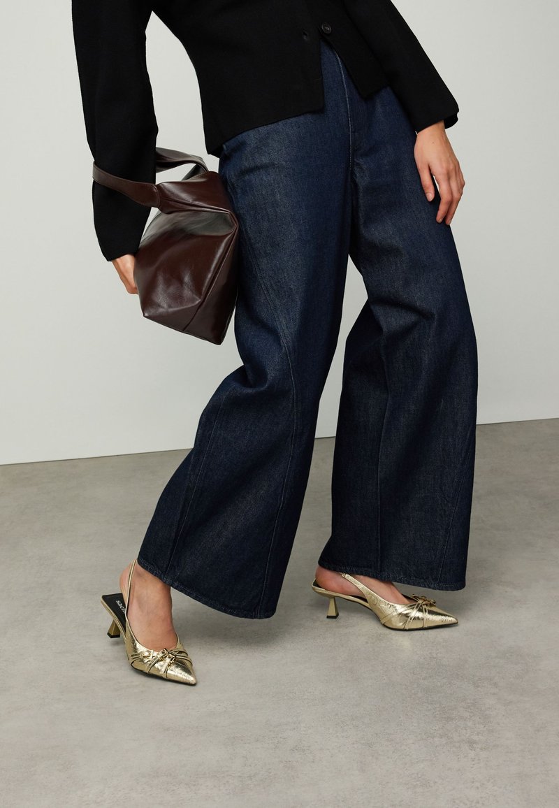 Pantalon large en denim foncé associé à des chaussures slingback métalliques dorées et un sac à main marron. Le tissu présente une texture lisse et un design structuré.