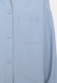 Světle modrá košile na knoflíky s náprsní kapsou s vyraženým názvem značky. Hladká textura a volný střih. Kulaté knoflíky ve stejné barvě.