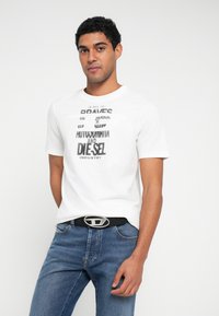 Tricou alb cu mâneci scurte și text grafic negru, asortat cu blugi albaștri și o curea neagră cu o cataramă ovală argintie. Design simplu.