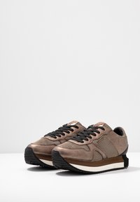 Brown sneaker featuring a textured fabric upper, black laces, rubber outsole, and a layered sole design with a slightly elevated heel.