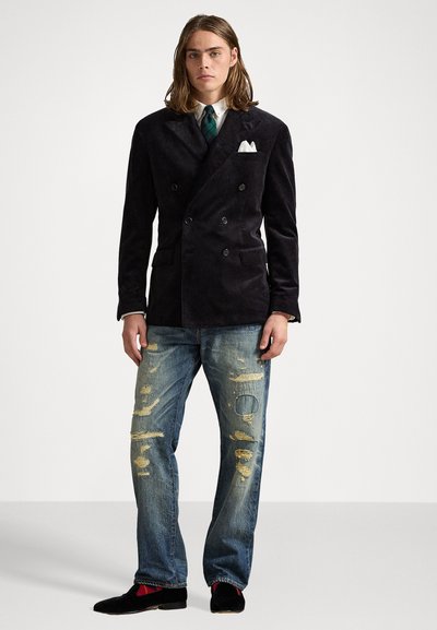 Man in zwarte dubbelgeboude blazer, witte shirt, groene geruite stropdas, versleten blauwe jeans, zwarte schoenen en rode gestreepte sokken, staand voor een witte achtergrond.