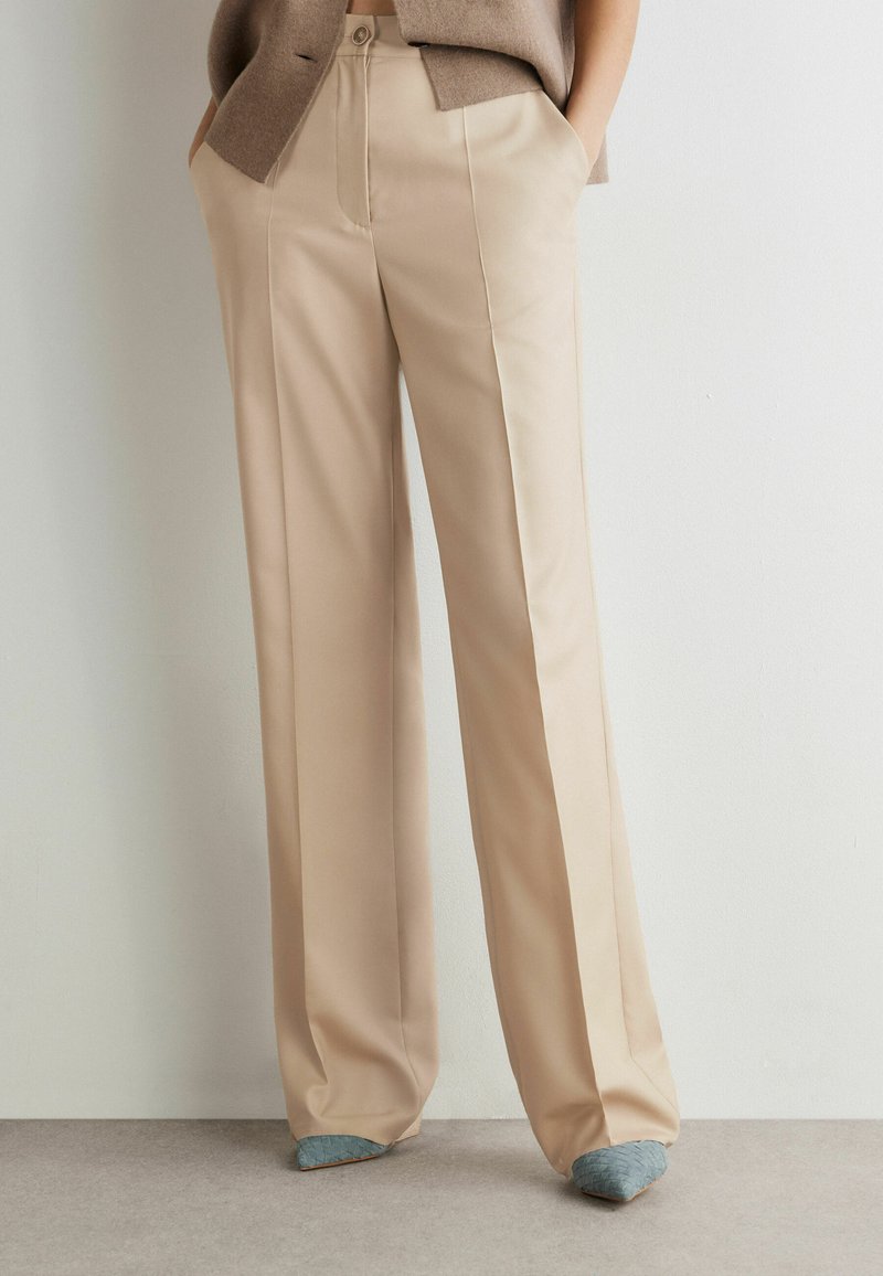 Personne portant un pantalon beige à jambes larges sur mesure avec des plis sur le devant, les mains dans les poches, associé à des talons bleus à bouts pointus.