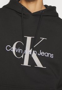 Sudadera negra con capucha hecha de una mezcla de algodón. Presenta un logotipo bordado en blanco "Calvin Klein Jeans" y cordones en la capucha.
