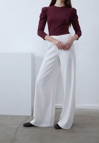 Burgunderfärgad långärmad topp med puffade axlar, parat med höga vita vida byxor och mörka platta skor på ett betonggolv.