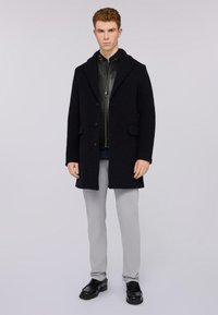 Cappotto in misto lana scuro con colletto a scialle, due bottoni e tasche laterali, indossato sopra una giacca nera con zip e pantaloni grigio chiaro. Scarpe nere.