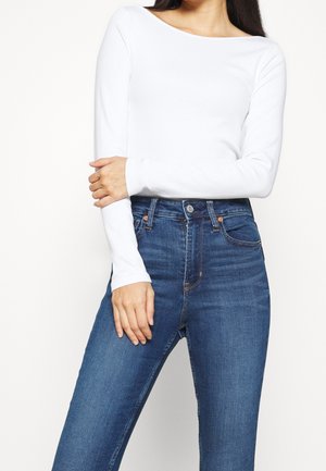 Frau trägt ein weißes langärmeliges Oberteil und hoch taillierte blaue Jeans, wobei ein Arm über den anderen verschränkt ist.