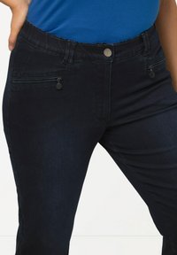 Jeans de mezclilla oscura con un corte ajustado, que presentan cremalleras en la parte delantera y una cinturilla robusta con un botón metálico. Textura suave y diseño clásico.