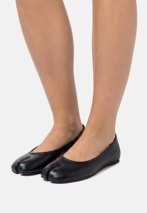 Pies con zapatos planos estilo tabi de cuero negro con diseño de punta dividida, sobre un fondo gris claro.