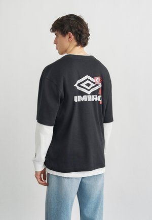 Joven con cabello rizado, vistiendo capas de mangas largas blancas y negras y jeans azul claro, mirando hacia la izquierda y mostrando un logo gráfico en la parte trasera.