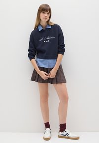 Sudadera navy con bordado blanco, superpuesta a una camisa a rayas azul claro, combinada con una falda de cuadros marrón con pliegues y zapatillas blancas.