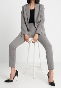 Szary w kratę garnitur składający się z podwójnie zapinanego żakietu i dopasowanych spodni, w połączeniu z czarnymi szpilkami.