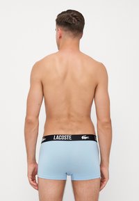Svetlo modri bokserji imajo črn pas z belim logotipom "LACOSTE" in aligatorjem. Imajo gladko teksturo in prilegan obliko.