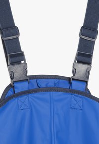 Μπλε αδιάβροχες φόρμες εργασίας με ρυθμιζόμενους ναυτικούς ιμάντες και πλαστικά εξαρτήματα, με λείες επιφάνειες και ενισχυμένες ραφές.