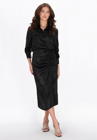 Vestido midi de satén negro con botones delanteros, mangas largas y detalle fruncido en la cintura. Combinado con sandalias plateadas de tiras.