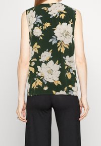 Ärmlös mörkgrön topp med stort vitt och gult blommönster, bärs med lösa svarta byxor, sedd från baksidan.