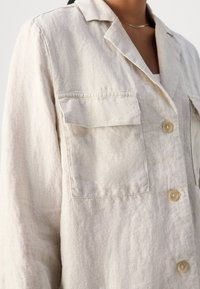 Giacca in lino beige chiaro con vestibilità comoda, colletto a rever, due tasche frontali e chiusura con bottoni, caratterizzata da una texture delicata.