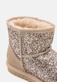 Glitterem pokrytá kotníková bota s hnědou semišovou základnou, měkkou kožešinovou podšívkou a béžovým prošívaným okrajem. Podešev je plochá s texturovaným gripem.