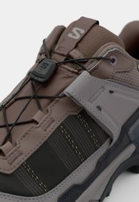 Trail running shoe featuring a mix of brown synthetic upper, mesh accents, flexible black sole, elastic laces, and a textured gray panel.