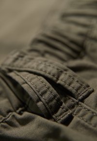 Tessuto khaki con una trama testurizzata, caratterizzato da sezioni arricciate e cuciture rinforzate. In evidenza una cinghia piegata con cuciture visibili.