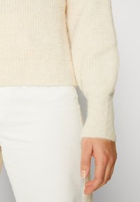 Gestructureerde crèmekleurige trui met ribbels aan de mouwen, zachte wolmix en een relaxte pasvorm. Draagt in combinatie met witte broeken, waarbij de mouwdetails goed zichtbaar zijn.