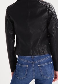 Giacca da motociclista in pelle nera con dettagli imbottiti sulle spalle, design aderente, colletto alto e retro accorciato. Abbinata a jeans di denim blu.