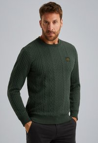 Groene gebreide trui met getextureerde kabelpatronen, ribbelboord bij de ronde hals en mouwen. Klein logo-embleem op de borst. Lange mouwen en een relaxte pasvorm.