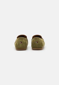Oliven grøn ruskinds slip-on sko med en brun gummisål og små logoer nær hælen. Blød tekstur, rundbakket design.