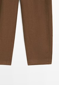 Pantalon brun à jambes droites avec un détail de couture visible près de l'ourlet inférieur sur un fond blanc uni.