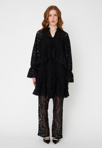 Rochie din dantelă neagră cu volane suprapuse și mâneci lungi, asociată cu pantaloni din dantelă transparentă asortați. Un design elegant și texturat cu modele florale.