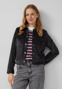 Zwarte corduroy jas met twee borstzakken, drukknoopsluitingen en een rechte snit, gecombineerd met een gestreept polo shirt en grijze spijkerbroek.