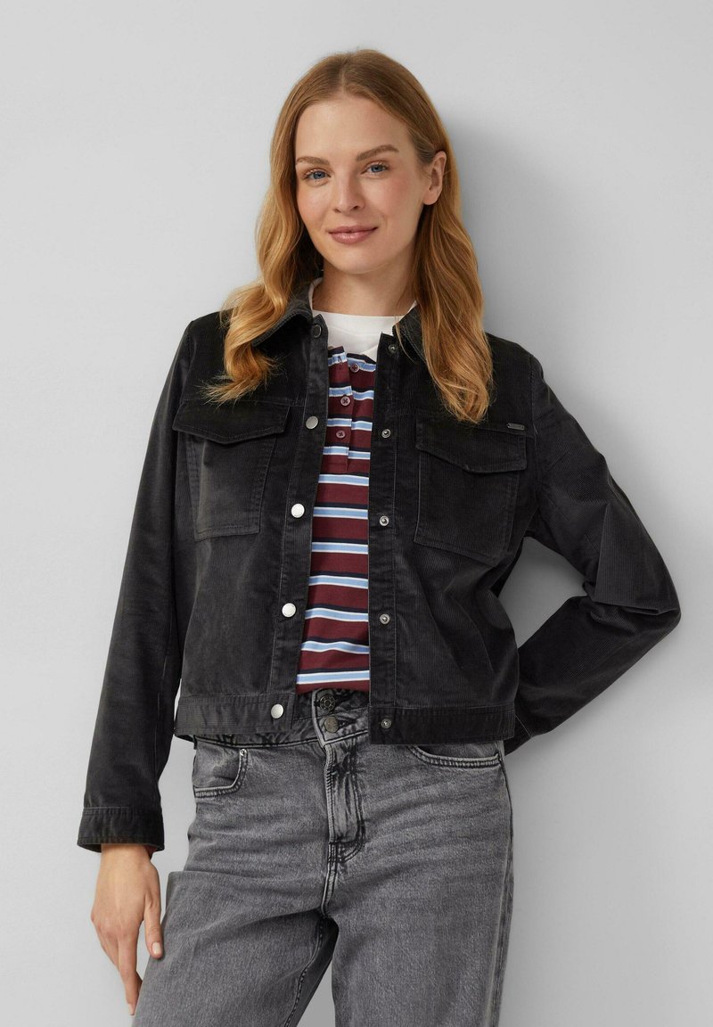 Zwarte corduroy jas met twee borstzakken, drukknoopsluitingen en een rechte snit, gecombineerd met een gestreept polo shirt en grijze spijkerbroek.