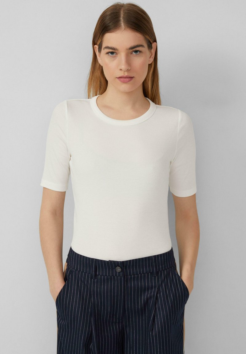 Haut blanc à manches courtes avec un col rond et une texture côtelée, associé à un pantalon bleu marine à rayures, les mains dans les poches, sur un fond neutre.