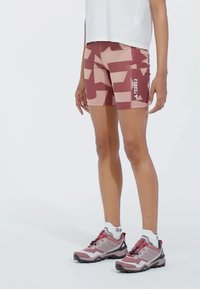 Terrex patterned shorts in shades of pink with a geometric design. Mid-thigh length, stretchy material, paired with pink and gray athletic shoes.